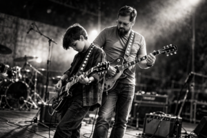 Pai e filho tocando guitarra juntos no palco, vistos de lado, em imagem em preto e branco, representando presença, orgulho e autonomia.